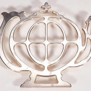Könnte beinhalten: Ein silberfarbener Metalluntersetzer in Form einer Teekanne. Das Design zeigt die ausgeschnittene Silhouette einer Teekanne mit einem zentralen, globusartigen Muster, einem geschwungenen Griff links, einem Ausguss rechts und einem Zierknauf.