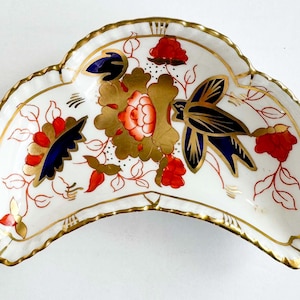 Può includere: Un piatto in porcellana a forma di mezzaluna con base bianca, caratterizzato da un motivo floreale centrale dorato, accenti rossi e blu scuro e l'illustrazione di un uccello stilizzato. I bordi sono smerlati e rifiniti in oro.