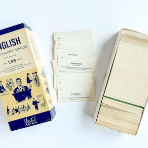 May include: A set of English Vocabulary Cards by Vis-Ed in a cream and navy blue box with vintage illustrations of people in professional settings. The set includes a stack of cream-coloured cards with printed words, definitions, and example sentences.