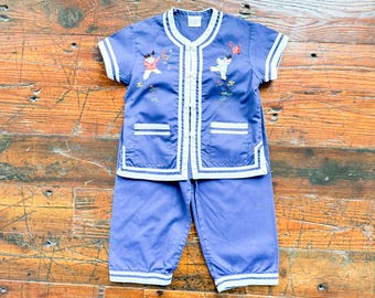 Puede incluir: Un conjunto de pijama azul bígaro de manga corta con cuello mao, ribetes blancos y cierres de alamares. La parte superior incluye dos bolsillos y bordados de niños jugando, junto con pantalones cortos a juego.