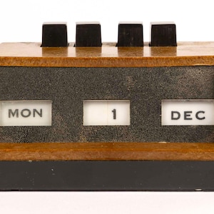 Puede incluir: Un calendario de escritorio vintage con marco de madera marrón y base negra. El calendario muestra "LUN 1 DIC" con botones negros en la parte superior para ajustes manuales. El diseño es retro y funcional.