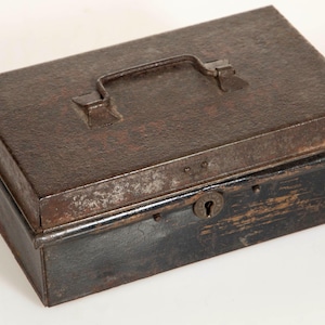 Puede incluir: Una caja de metal antigua, rectangular, con asa y cerradura. La caja es principalmente marrón oscuro con una superficie texturizada, y la parte inferior está pintada de negro. El asa es del mismo metal.
