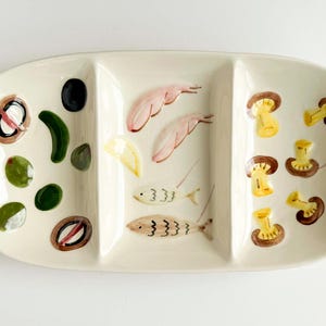 May include: Cream-colored, oval-shaped serving dish with three compartments. The dish is decorated with painted olives, shrimp, fish, and mushrooms. The dish is a decorative piece for serving appetizers or snacks.