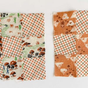 May include: Two square fabric quilt blocks composed of nine smaller patches each. The left block features a mix of orange grid patterns and a cream-colored print with small houses, trees, and figures. The right block combines orange grid patterns with a solid orange print featuring white floral and geometric shapes.