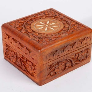 May include: A square wooden box with a hinged lid featuring intricate floral carvings along the sides and top. The center of the lid displays a circular inlay with a light-colored, star-shaped pattern surrounded by carved leaves and flowers.
