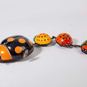 May include: A wind-up tin toy featuring a large black ladybug with orange spots connected to three smaller ladybugs in yellow, orange, and green. Each ladybug has painted eyes, antennae with red tips, and a metal winding key on the side of the largest.