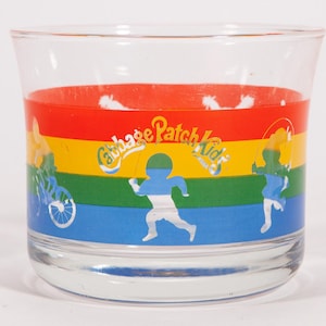 May include: A clear glass tumbler with a colorful design featuring the text "Cabbage Patch Kids." The design includes a rainbow of colors and cartoon figures of children playing. The glass is cylindrical with a slightly flared top.
