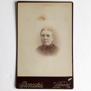 Puede incluir: Fotografía de retrato en tonos sepia de una persona con el pelo recogido, vestida con una prenda oscura. La fotografía está montada en una tarjeta rectangular con el nombre "Thevaskis" y "Varzleton, PA" impresos en la parte inferior.