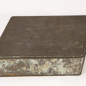 May include: A rectangular metal box with a lid. The box has a distressed, aged appearance with a dark grey colour and areas of rust and discolouration. The box is likely vintage or antique.