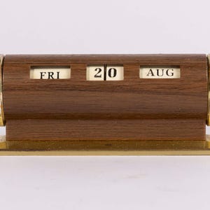 May include: A horizontal desk calendar with a dark brown wood-grain finish, featuring three rectangular windows displaying the text FRI, 20, and AUG. The cylindrical body is capped with metallic gold ends and rests on a rectangular gold base.