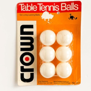 Peut inclure: Un paquet de six balles de tennis de table. L'emballage est orange et blanc, avec les mots "Table Tennis Balls" en haut. Le nom de la marque "Crown" est imprimé verticalement sur le côté gauche. Les balles sont blanches et dans un blister transparent.