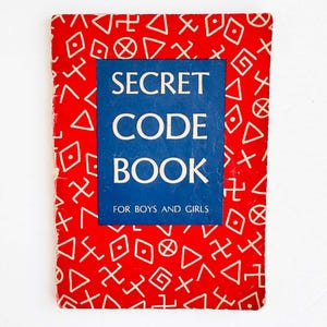 Peut inclure: Un livre rectangulaire avec une couverture rouge vif ornée d'un motif répétitif de symboles géométriques blancs. Un panneau rectangulaire bleu central affiche le texte blanc SECRET CODE BOOK suivi du texte FOR BOYS AND GIRLS.