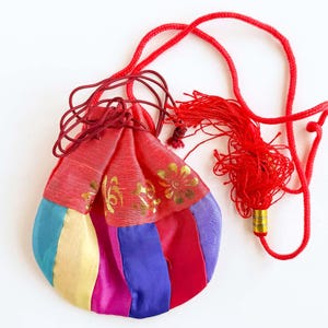 May include: A colorful, patchwork pouch with a red drawstring and tassel. The pouch features panels of blue, yellow, pink, and purple fabric, with a red top section adorned with gold floral designs. The tassel and drawstring are a vibrant red.