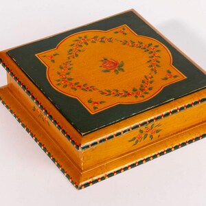 May include: A square wooden box with a hinged lid. The box is painted with a mustard yellow base and a dark green lid. The lid features a floral design with red and orange accents. The edges are decorated with small red and black dots.