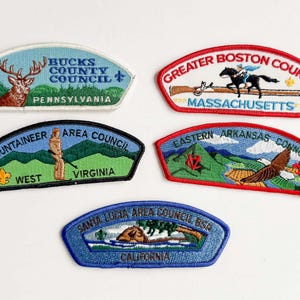 May include: Five embroidered patches featuring various designs and text. The patches include a deer, a horse and rider, a figure with a rifle, an eagle, and a coastal scene. Each patch has text indicating a council and state.