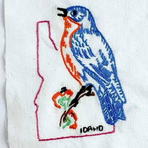 May include: A white fabric patch featuring an embroidered blue bird with an orange chest perched on a branch. The design includes a pink outline of the state of Idaho, green floral accents, and the word IDAHO stitched in black thread.