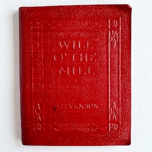May include: A small, red book with embossed text and decorative borders. The title "Will O' the Mill" is visible, along with the author's name, Stevenson. The book has a textured cover and appears to be vintage.