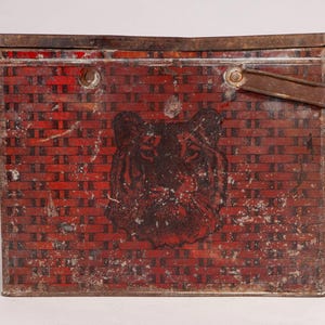 May include: A rectangular metal tin with a weathered red and black woven basket pattern. The front features a prominent black tiger head illustration. The container has a hinged lid and a single metal handle attached to the top right side.