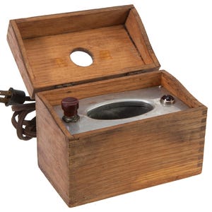 May include: An antique wooden box with a hinged lid, featuring a metal interior and two red knobs. A power cord with a two-prong plug extends from the side. The box is made of light brown wood and has a circular opening in the lid.
