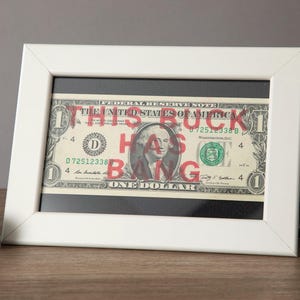 May include: A framed one-dollar bill with the words "THIS BUCK HAS BANG" in red over the original text. The white frame sits on a wooden surface, with a film roll and sunglasses in the background.