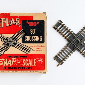 May include: An Atlas HO scale 90-degree crossing track and its packaging. The box is tan and red with the Atlas logo and product information. The track is black with brass-colored rails, designed for HO scale model trains.