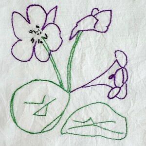 May include: A piece of off-white fabric featuring a hand-embroidered design of flowers and leaves. The floral petals are outlined in purple thread with black center details, while the stems and rounded leaves are stitched in green thread.