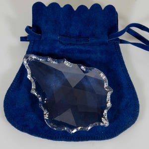 May include: A clear, faceted crystal decorative piece with a scalloped edge rests on a dark blue velvet pouch. The pouch has a drawstring closure and a scalloped top edge. The crystal is a unique shape.