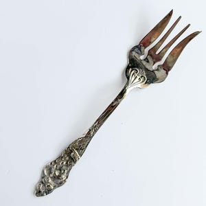 Op de afbeelding: Een zilverkleurige metalen vork met een decoratief handvat voorzien van reliëfpatronen van bloemen en bladeren. De tanden van de vork zijn breed en gebogen en vertonen tekenen van aanslag en oxidatie op het metalen oppervlak.