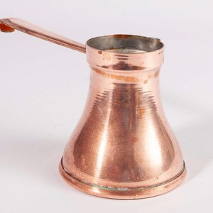 May include: A copper Turkish coffee pot with a wide, rounded base that tapers toward a narrow neck and a flared rim. The pot features a long, flat, angled handle extending from the side and a small pouring spout integrated into the top rim.