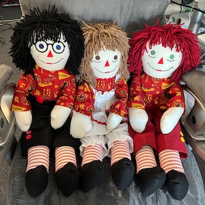 May include: Three handmade rag dolls dressed in red and black Gryffindor robes with yellow accents. The dolls have black, brown, and red hair and are sitting on a gray chair.