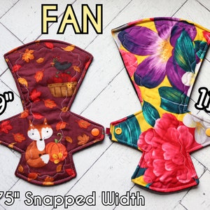 May include: Two cloth menstrual pads labeled FAN. The left pad is 9 inches long with a maroon background, autumn leaves, a fox, and a pumpkin. The right pad is 11 inches long with a yellow background and large purple and red flowers. Both have a 2.75 inch snapped width.
