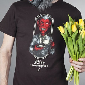 May include: A person wearing a brown t-shirt with a graphic of a red devil holding a teacup. The text "Nice to meet you" is printed below the graphic. The person is also wearing a gray wolf mask and holding a bouquet of yellow tulips.
