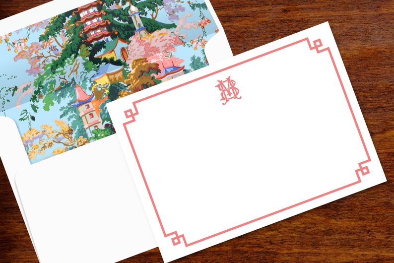 Pode incluir: Conjunto de papelaria branco com borda e monograma rosa. O interior do envelope apresenta uma paisagem colorida de inspira&ccedil;&atilde;o asi&aacute;tica com pagodes e &aacute;rvores. A papelaria est&aacute; sobre uma superf&iacute;cie de madeira escura.