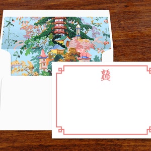 Pode incluir: Um conjunto de papelaria com um cart&atilde;o branco com uma borda coral e um envelope a condizer. O interior do envelope apresenta uma paisagem colorida de inspira&ccedil;&atilde;o asi&aacute;tica com pagodes e &aacute;rvores. O cart&atilde;o tem um monograma no canto superior direito.