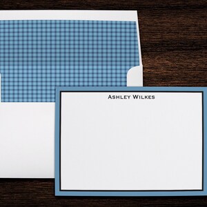 May include: A white envelope with a blue and white checked liner. A blue and white card with a black border and the text "ASHLEY WILKES" is next to the envelope.