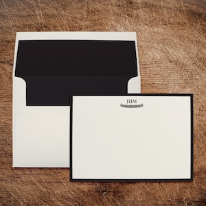 May include: A white envelope with a black inner flap and a black border around the edge. The envelope is lying on a wooden surface. A white card with a black border is lying on top of the envelope. The card has the initials "JHM" and a canoe graphic printed on it.