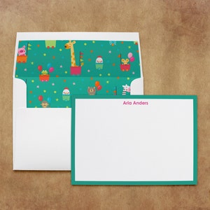 May include: A white envelope with a green and white patterned flap. The flap features a variety of animals in colorful wagons. The envelope is open to reveal a white card with a teal border and the name "Aria Anders" printed in pink.