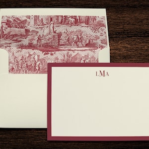 May include: A white envelope with a red and white patterned flap. The envelope is closed and has a white card inside with a red border and the monogram "LMA" printed in red.