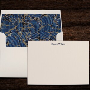 May include: A white envelope with a blue and gold marbled pattern on the flap. The envelope is open and a white card with the name "Beaux Wilkes" printed in blue ink is inside.
