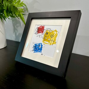 May include: A framed drawing of three cartoon faces in a black frame. The faces are drawn in red, yellow, and blue.