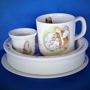 Könnte beinhalten: Ein weißes Porzellan-Teetassen- und Untertassen-Set mit einer Beatrix Potter-Illustration von zwei Kindern und einer Katze. Die Tasse hat einen Henkel und die Untertasse einen gewellten Rand.