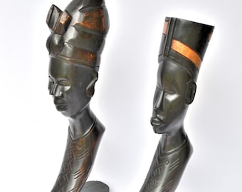 May include: Two dark brown wooden sculptures of human busts. Each has a tall, ornate headdress with copper-coloured accents. The figures have detailed facial features and are mounted on rectangular bases. These sculptures are likely decorative art.