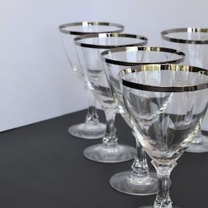 Peut inclure: Cinq verres à vin en verre transparent avec une bordure argentée. Les verres sont disposés en rangée sur une surface noire.