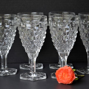 May include: Six clear glass goblets with a diamond pattern on the bowl and a hexagonal base. The goblets are arranged in a row on a black surface with a single orange rose in front of them.
