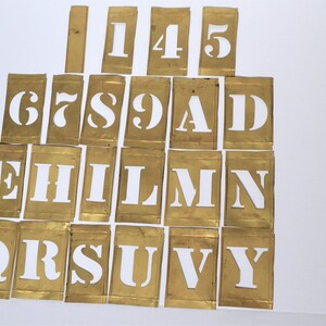 May include: A set of brass stencils with white letters and numbers, including the letters E, H, I, L, M, N, Q, R, S, U, V, Y and the numbers 1, 4, 5, 6, 7, 8, 9, A, and D.