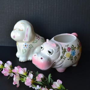 May include: Two ceramic figurines, one a white dog with pink ears and floral accents, the other a pink hippopotamus with floral accents. Both figurines are painted with pink, blue, and yellow flowers.