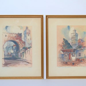 May include: Two framed watercolor paintings depicting scenes from a city in Morocco. The left painting shows a street with an archway and buildings. The right painting shows a building with a tower and a small house.