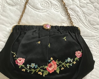 Pode incluir: Bolsa de noite de cetim preto com alça de corrente dourada. A bolsa apresenta um design de bordado floral em rosa, azul e verde. Um fecho decorativo com uma pedra rosa e dourada garante o fecho da bolsa.
