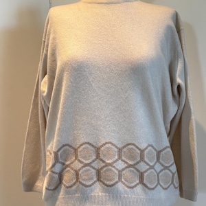 May include: A cream-coloured sweater with a crew neck and long sleeves. The bottom of the sweater features a geometric pattern of interlocking hexagons in a lighter cream colour. The sweater is displayed on a mannequin.