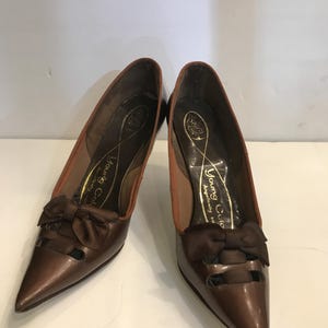 May include: A pair of brown leather pumps with pointed toes and small bows. The shoes have a glossy finish and a small heel. The insoles are tan with gold text that reads "Young Colony, Angelically so".